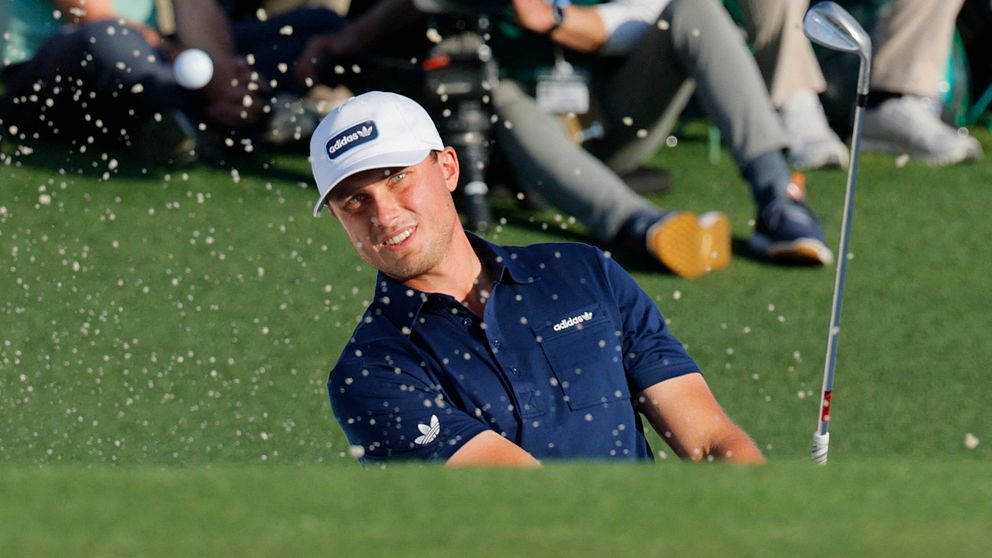 Golfspelare slår ett slag ur en sandbunker, sandpartiklar sprids i luften.