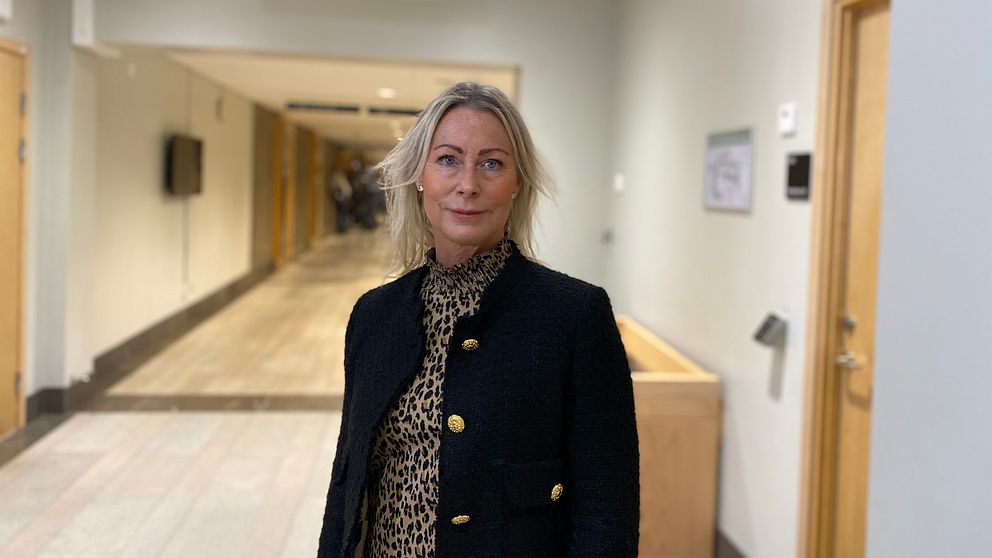 En person står i en korridor med neutrala väggar och belysning. Personen bär en svart kavaj och en blus med leopardmönster. I bakgrunden syns en annan person och en dörr som leder till en toalett, markerad med skyltar.