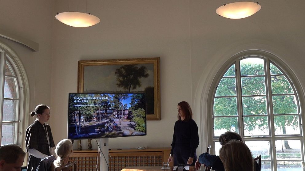 En presentation pågår i ett ljust rum med stora fönster. En kvinna står framför en skärmtv som visar en bild av en park. En annan kvinna, till vänster, verkar förbereda sig för att tala. Rummet innehåller också konstverk och möbler i trä. Deltagarna i rummet är fokuserade på presentationen.