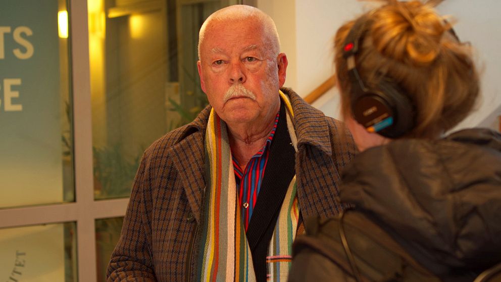 Man med vitt kort hår iklädd en rutig rock och en randig halsduk. Mannen tittar snett förbi kameran. I förgrunden en kvinna med hår i knut och hörlurar, svart jacka.