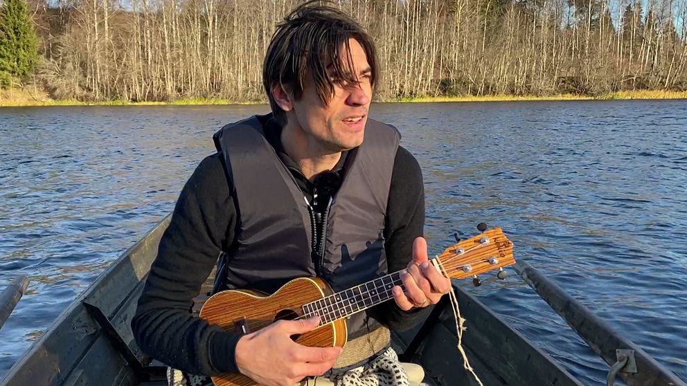 Gustaf Nohrén sitter i en båt på en älv och spelar på en ukulele. Runt omkring finns skog med lövträd, och det är en klar dag med soligt väder. Vattnet speglar omgivningarna.
