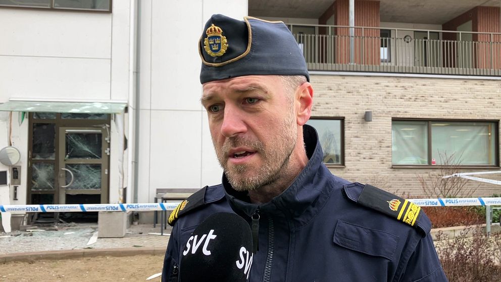 Anders Persson, Falkenbergspolisens operativa samordnare, i polisuniform står framför en byggnad med en trasig dörr och fönster, omgivet av avspärrningar.
