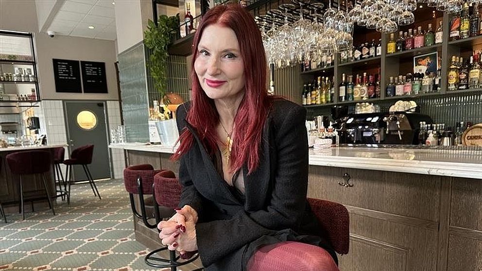 En person sitter på en stol i en stilfull bar med en modern inredning. Bakgrunden visar en välfylld barhylla med olika drycker och glas upphängda över bänken. Inredningen är elegant med gröna växter och en mönstrad matta som pryder golvet. Personen är klädd i en svart kavaj och röda leggings, vilket ger en modebetonad känsla.
