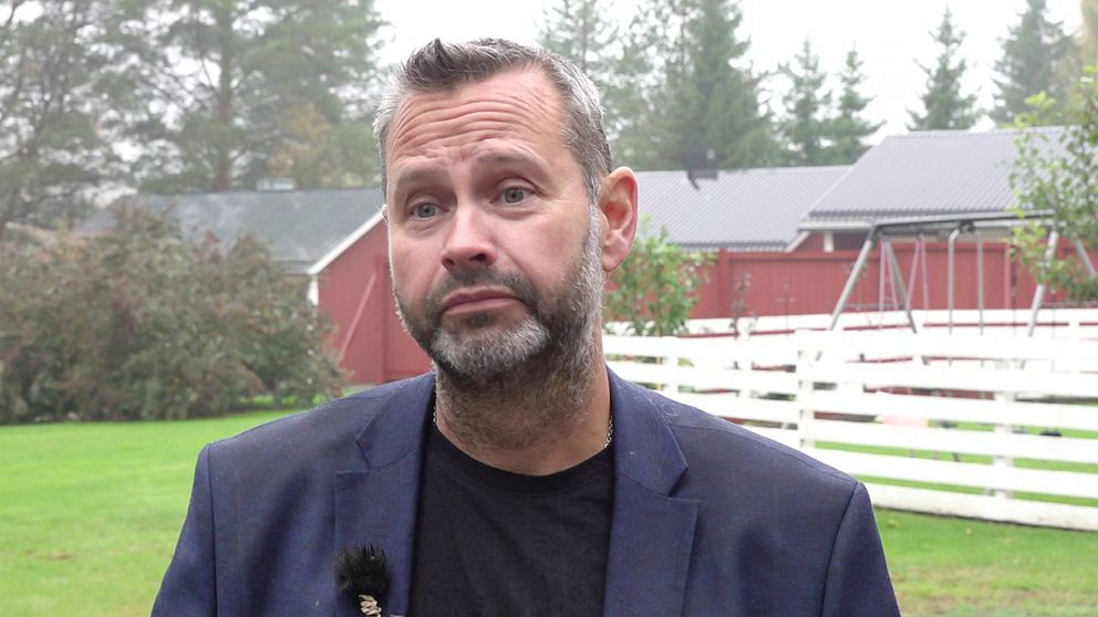 Mattias Riegertzon står framför en grön gräsmatta med träd och en röd byggnad i bakgrunden. Han  bär en mörk jacka och har en mikrofon fäst på kläderna. Det är en dimmig dag, vilket ger en dämpad atmosfär.