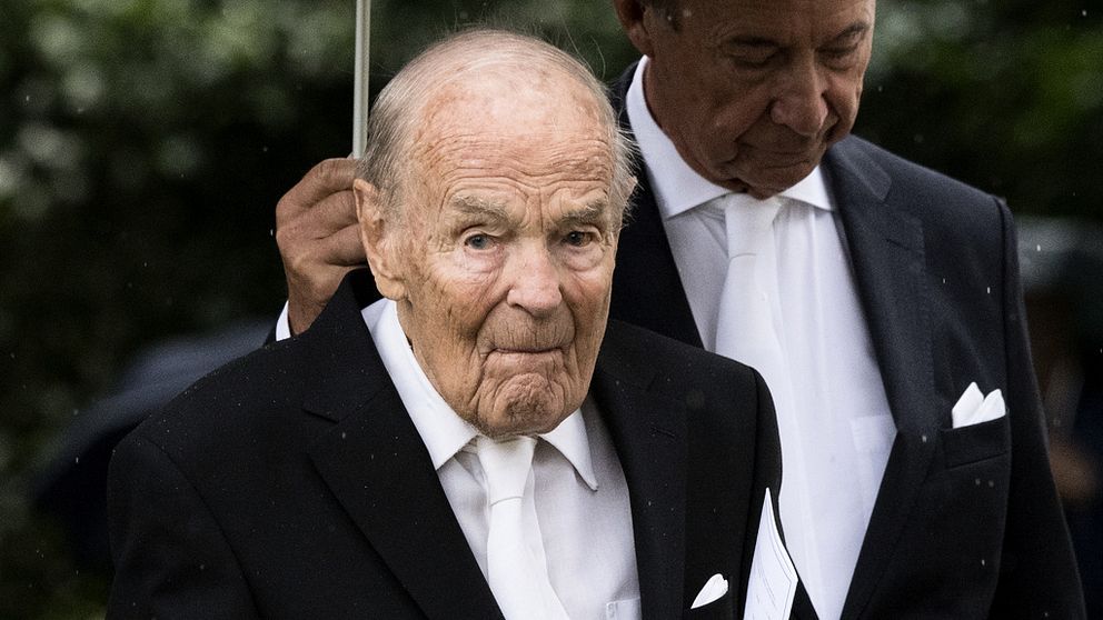 Hans Gabriel Trolle Wachtmeister i svart kostym, vit skjorta och vit slips. En man bakom honom håller upp ett paraply över honom.