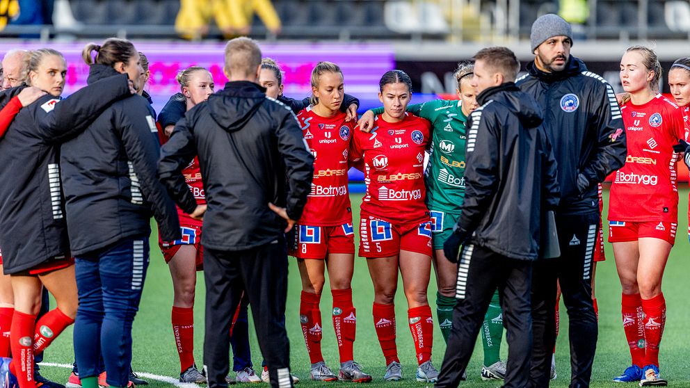 Fotbollsspelare i röda matchkläder och tränare i svarta jackor står i en ring och har ett lagmöte på en fotbollsplan.