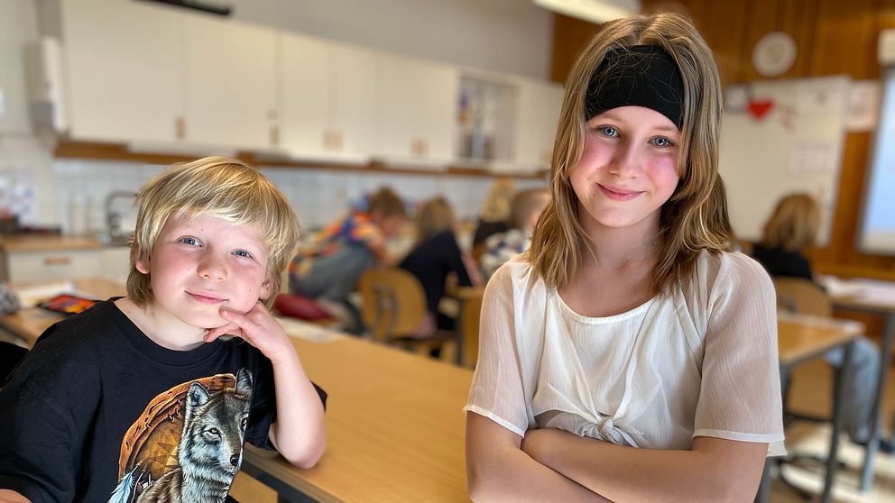 Två elever i klassrum. Vända mot kameran. En pojk som sitter i sin bänk och en flicka som står intill honom.