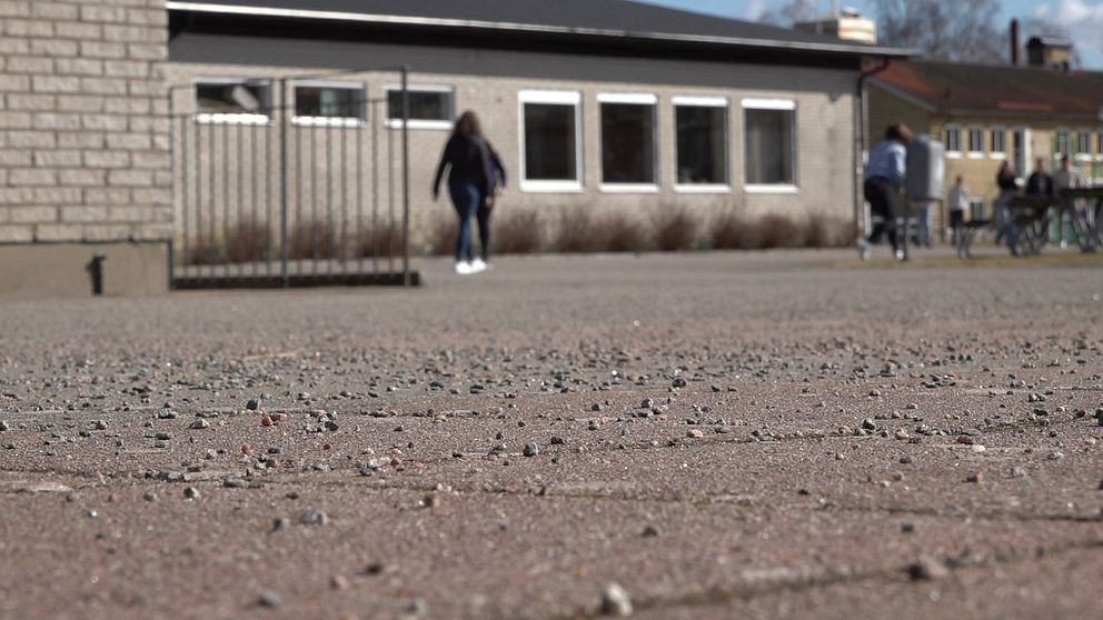 En nära vy av en gård med grus och plattor i förgrunden. I bakgrunden syns Apelskolan. Två personer går på gården, vilket ger en känsla av aktivitet i miljön.