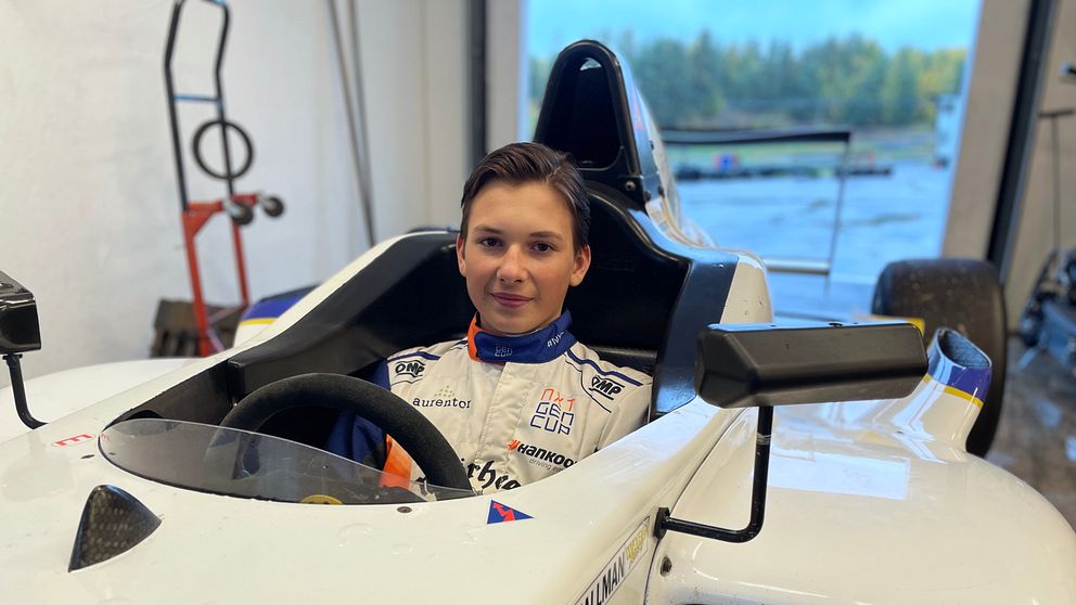 En förare sitter i en racingbil i ett garage. Bilen har en vit lackering med blå och gula detaljer. Bakgrunden visar delar av garaget och ett fönster med utsikt över en grönskande miljö. Föraren bär en overall med sponsormärken och är fastspänd i sätet.