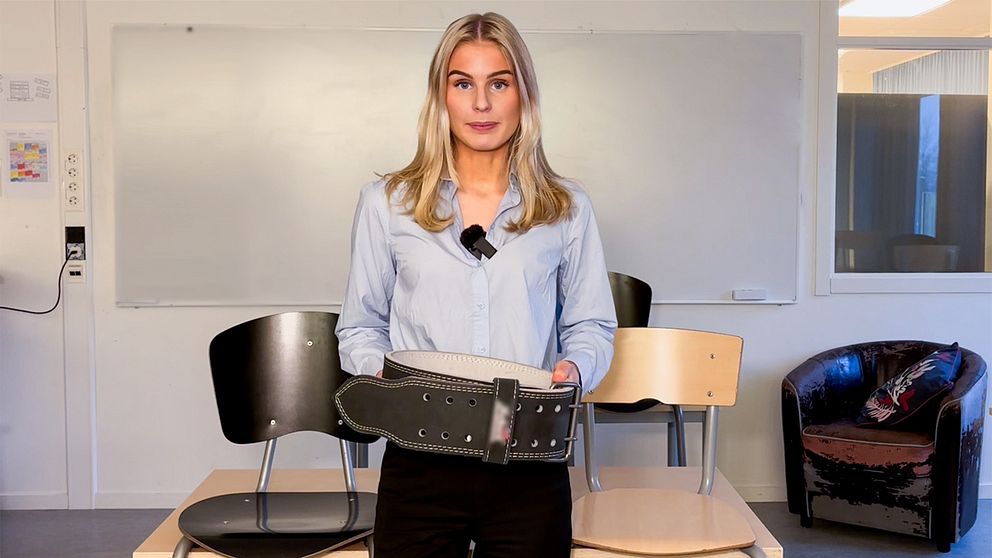 Alva Olsson, reporter på SVT, står i ett klassrum och håller i ett tyngdlyftarbälte.