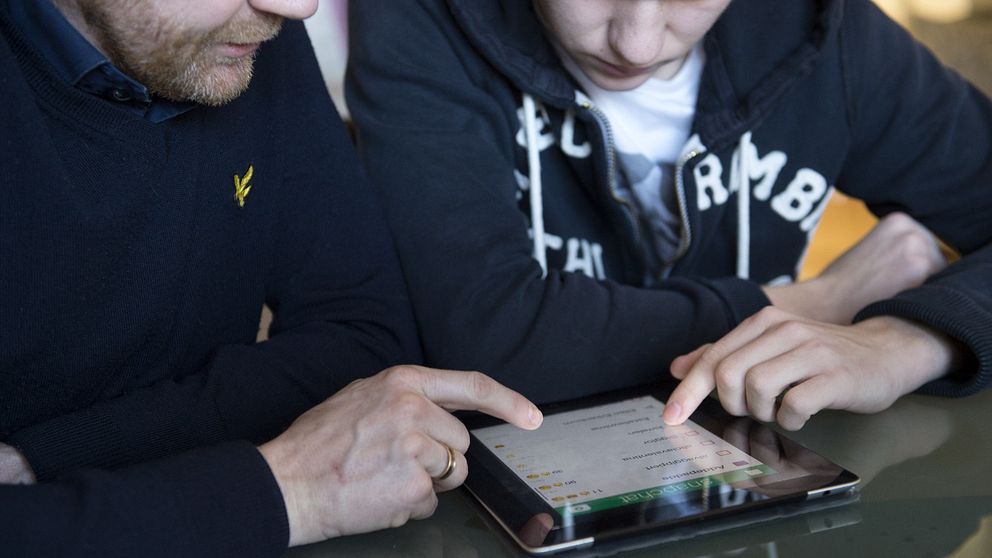 En man och en tonåring sitter vid ett bord och tittar på en surfplatta. Båda håller pekfingrarna över skärmen, där de interagerar med innehållet. Mannen har på sig en mörktröja och tonåringen en grå hoodie. Fokus ligger på deras händer och surfplattan i mitten av bilden.