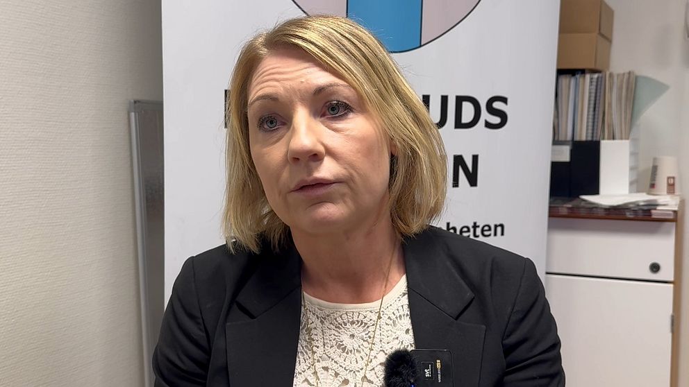 En person i en svart kavaj sitter framför en bakgrund med en logotyp som innehåller blå och rosa element. Personen har på sig en ljus blus och det finns dokument och en skrivbordssak synliga i bakgrunden.