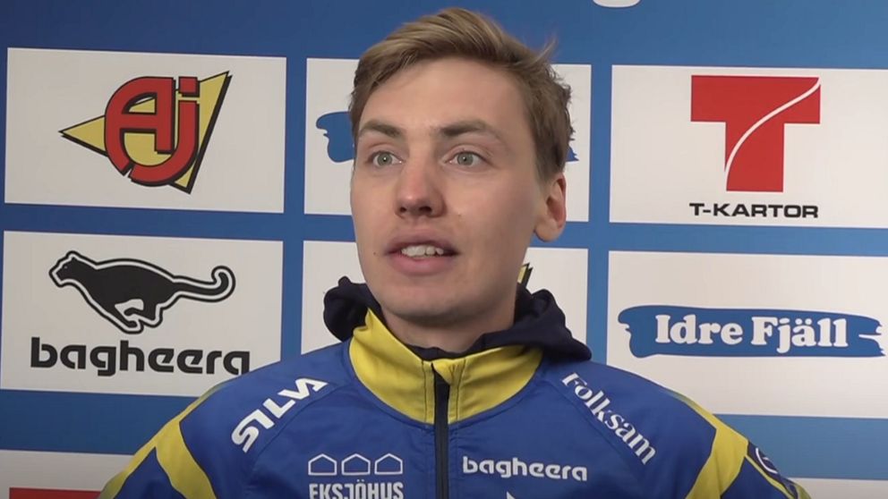 Person i blågul sportjacka framför sponsorvägg med logotyper.