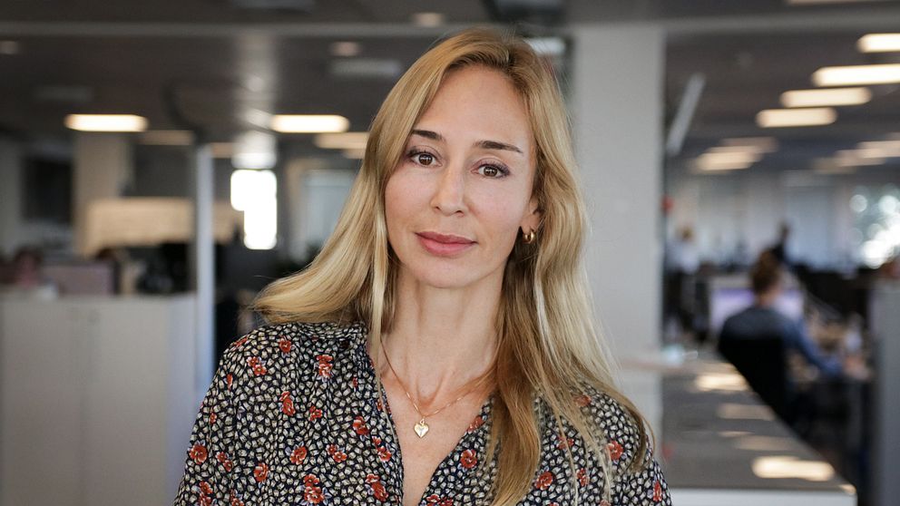 Kvinna med blont hår står på en kontorsarbetsplats med skrivbord och datorer i bakgrunden.