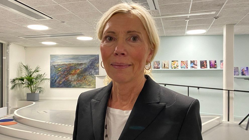 En kvinna i mörk kavaj står i inomhusmiljö med ett konstverk i bakgrunden. Rummet har skarpa, moderna linjer och är inrett med växter samt en hylla fylld med böcker eller tidskrifter på en vägg.
