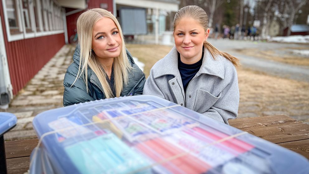 Två unga kvinnor ler bakom en plastlåda fylld med förnödenheter.