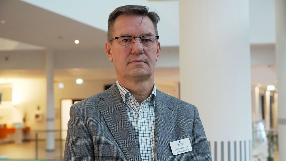 En person iklädd en grå kavaj med en namnbricka står inomhus i en modern byggnad med vit interiör och diskreta belysningsarmaturer i bakgrunden.