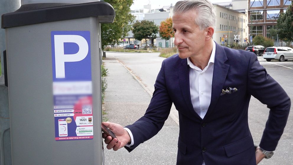 Kemal Hoso (S), ordförande i exploateringsnämnden i Örebro står nära en parkeringsautomat med en P-skylt på framsidan. Han håller en nyckel eller kort i handen och ser ut att interagera med automaten. I bakgrunden syns en grönskande gata med byggnader och träd.