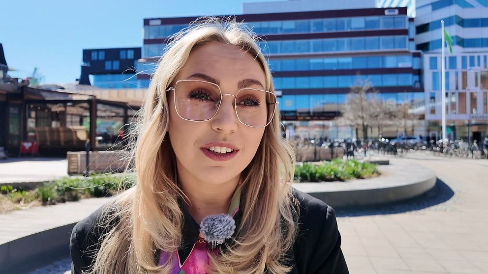 Kvinna med långt blont hår står utomhus i en stadsmiljö med moderna byggnader i bakgrunden.