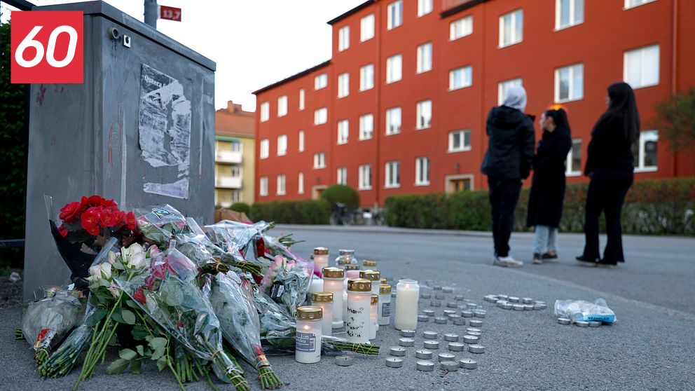 Blommor och tända ljus på trottoar, tre personer samlade i bakgrunden vid rött bostadshus.
