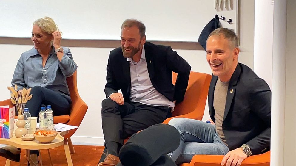 Tre personer sitter på orange fåtöljer under en diskussion eller paneldebatt. Det finns en liten kaffebord med en skål snacks och två flaskor med vatten i förgrunden.