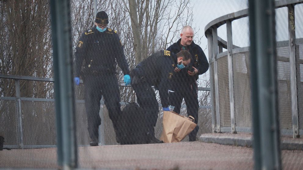 Poliser i skyddshandskar bär en kropp insvept i en brun väska över en gångbro.