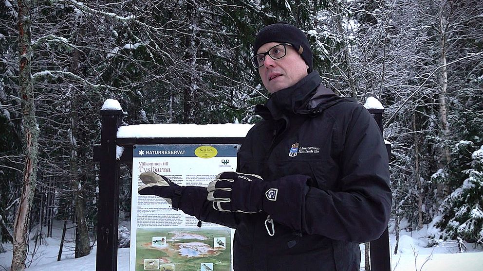 Mattias Västfelt som är chef Naturförvaltningsfunktionen står framför en informationsskylt i en snötäckt skog. Skyltens text och bilder handlar om området och dess natur, medan personen gestikulerar. Träden runt omkring är täckta av snö, vilket skapar en kylig och lugn atmosfär.