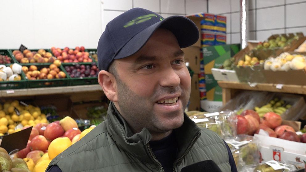 Person i butik med hyllor fyllda med frukt och grönsaker i bakgrunden.