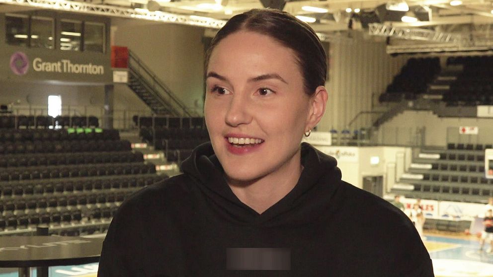 Ung kvinna i svart tröja står i en tom idrottsarena, läktare och basketplan syns i bakgrunden.