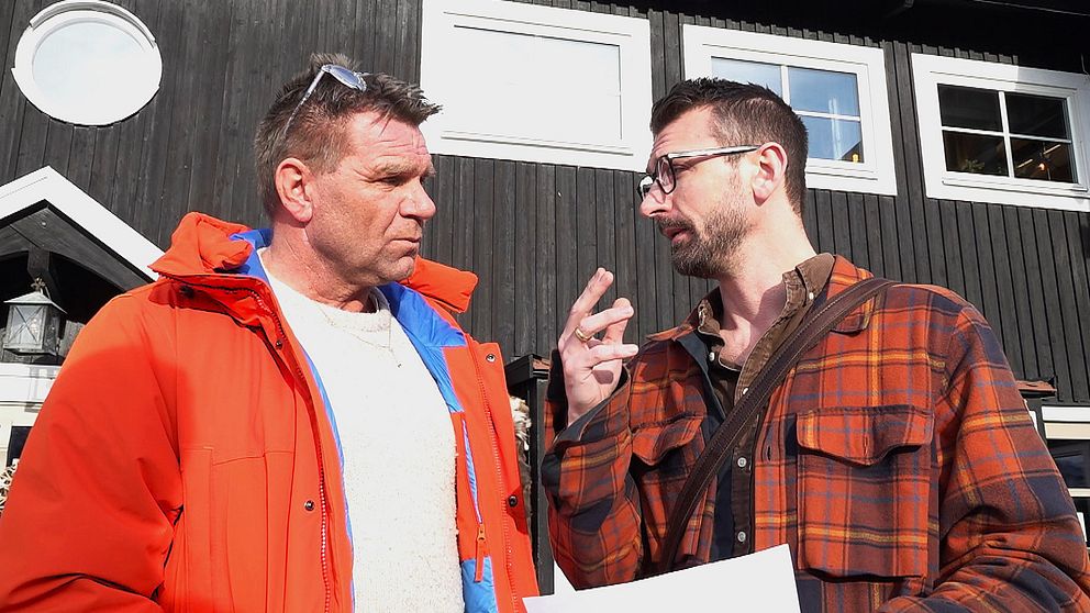 Två personer diskuterar utomhus framför ett mörkt trähus i solen.