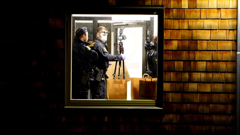 Poliser iklädda skyddsutrustning står inne i ett rum som syns genom ett fönster. De håller i en kamera på ett stativ och verkar förbereda sig för att dokumentera en scen, med en brun väska placerad på golvet. Bakgrunden består av en mörk vägg med trätak.