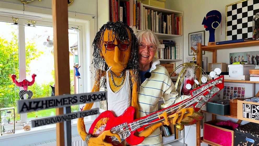 En person står bredvid en livfull orange docka som håller en röd gitarr, inuti ett rum fyllt med böcker och färgglada konstverk. I förgrunden syns en skylt med texten ”ALZHEIMERS SJUKDOM”. Rummet har stora fönster som släpper in naturligt ljus och ger en kreativ atmosfär.