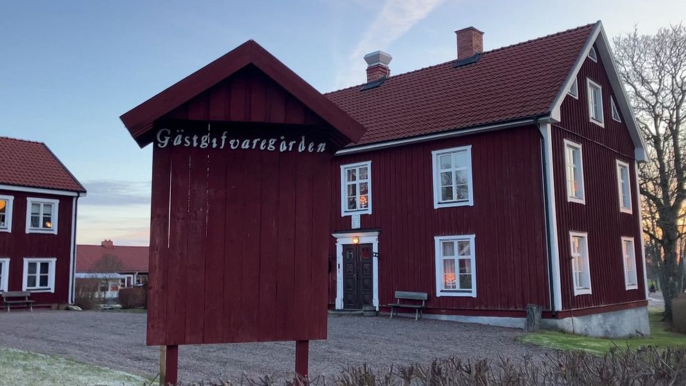 En röd timrad byggnad med vita fönster, tydligt markerad som ”Gästgivargården” på en skylt framför huset. Byggnaden har ett traditionellt utseende med två skorstenar och ligger i en lantlig miljö.