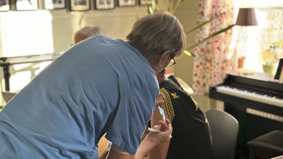 En person får en vaccination i en inomhusmiljö med en lugn atmosfär. Den som administrerar vaccinet står bakom och använder en spruta, medan den vaccinerade personen sitter ner och har på sig en svart t-shirt. I bakgrunden syns en piano samt fotografer på väggarna.