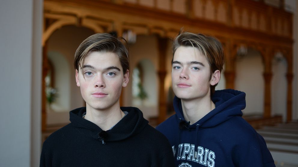 Två personer står i en byggnad med inredning som tyder på en historisk eller religiös miljö, med trädetaljer och valv i bakgrunden.
