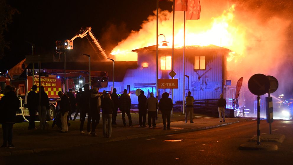 En nattlig scen där flera personer observerar en byggnad som brinner intensivt. Röd och orange eldslåga syns på taket och en brandbil är parkerad i närheten. Brandmän arbetar för att släcka elden.