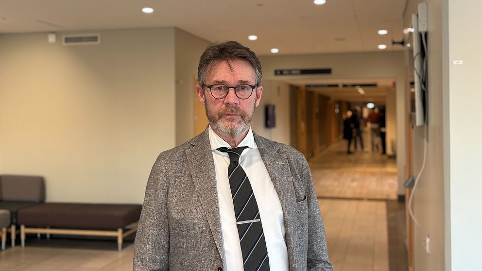 Man i kostym står i en korridor med flera dörrar och människor i bakgrunden.