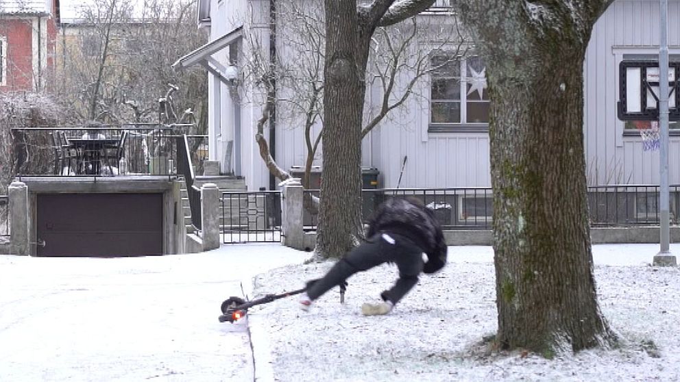 En person i svart kläder faller framåt på en snötäckt gård, nära en stor trädbas, med en bostad i bakgrunden och en basketkorg. Området ser kallt och vintrigt ut.