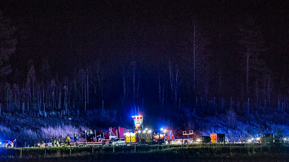 Bild av en räddningsoperation med flera nödbilar och ljus i en skogsmiljö. Personal arbetar nära en stor maskin, omgiven av blå och orangea varningsljus. Träd syns i bakgrunden, och stämningen antyder en intensiv insats.