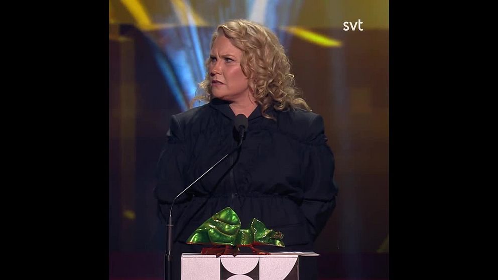 En person på scenen som står vid en mikrofon och håller i en festlig present insvept i grönt och guld. Bakgrunden har en färgglad belysning som skapar en festlig atmosfär.