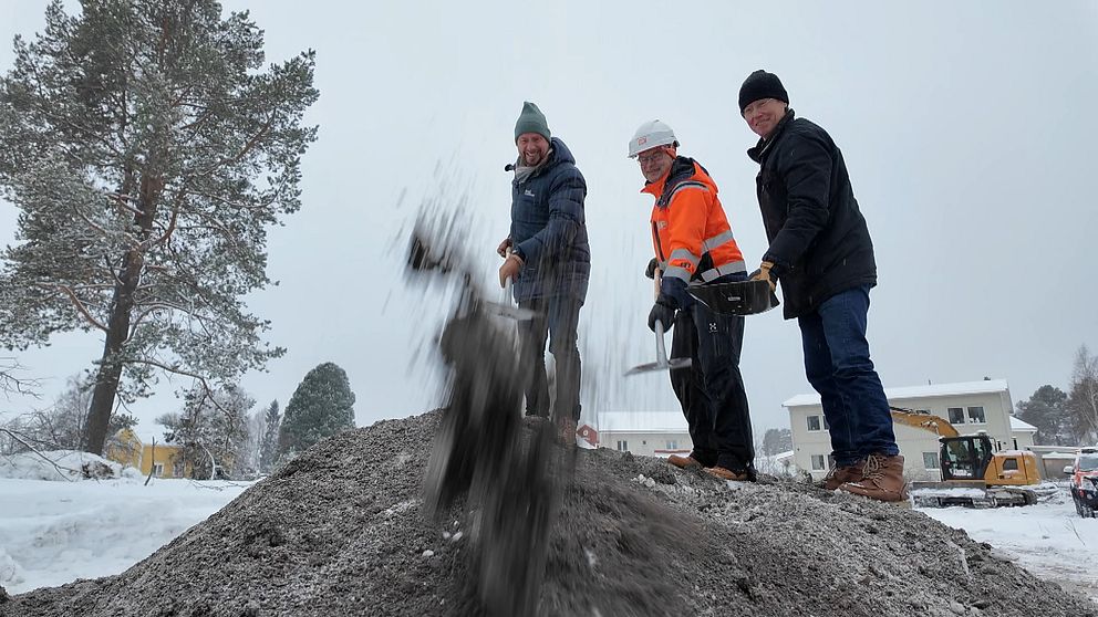 Tre personer kastar jord eller grus med spadar på en vinterlig byggarbetsplats. En man i orange skyddsväst står bredvid en person i mörk jacka medan en tredje person är på väg att kasta materialet. Bakgrunden visar träd och några byggnader.