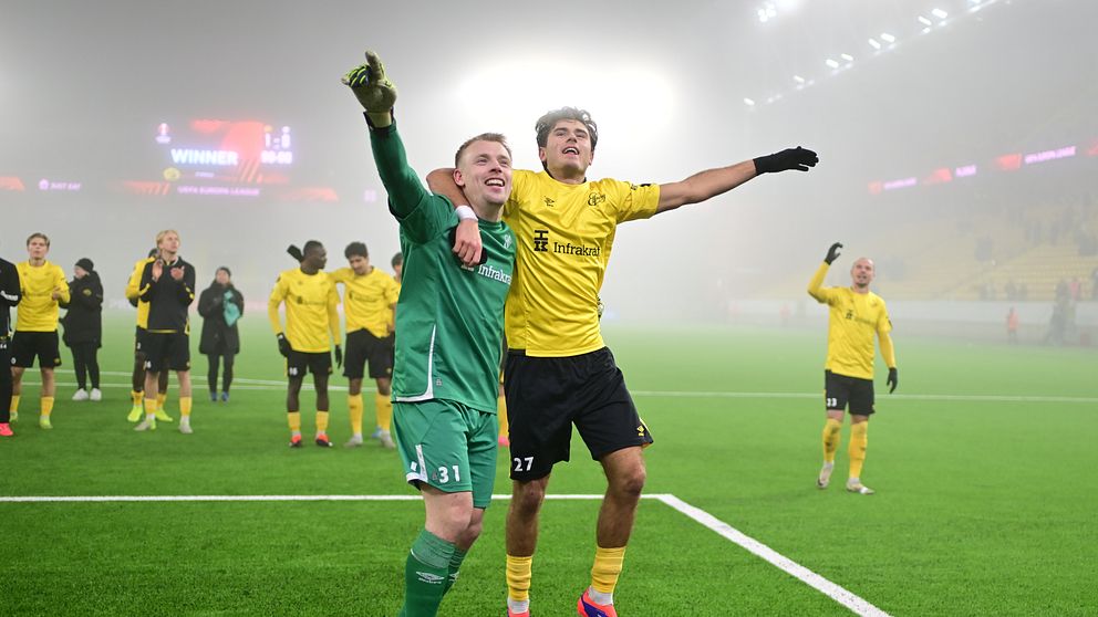 Två Elfsborgsspelare firar en seger på fotbollsplanen, omgivna av dimma. Den ena spelaren, i grön målvaktsdräkt, håller i den andra spelaren, som bär en gul tröja. I bakgrunden syns lagkamrater som deltar i firandet.