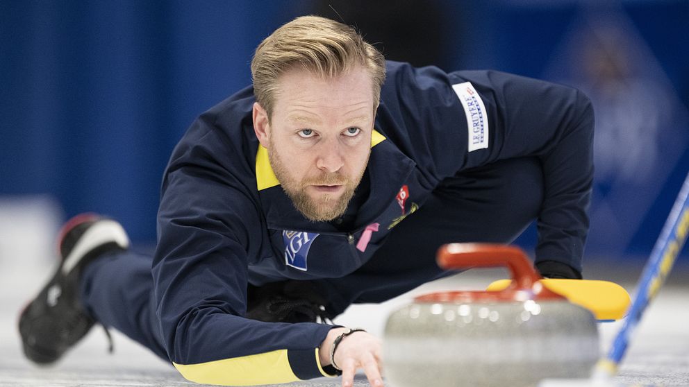 En curlingspelare ligger på knä på isen, sträcker sig fram mot en curlingsten med en snabb rörelse. Denna spelare bär en mörk jacka med gula detaljer och visar en koncentrerad hållning, med fokus på spelet. Curlingstenen ligger nära spelaren, på väg mot målet. Bakgrunden är suddig, vilket framhäver spelaren och stenen.
