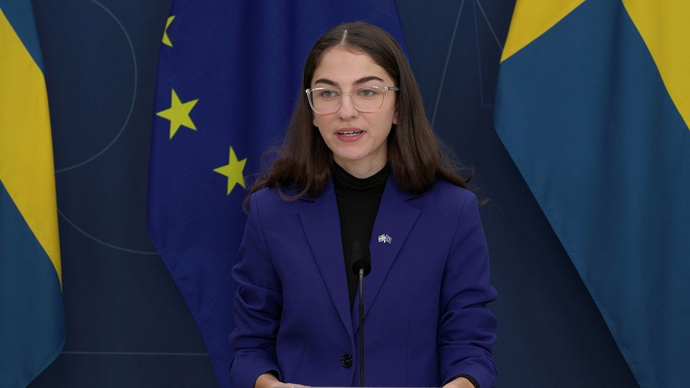 En person står vid ett podium under en presskonferens, klädd i en blå kavaj. Bakgrunden består av svenska och europeiska flaggor.