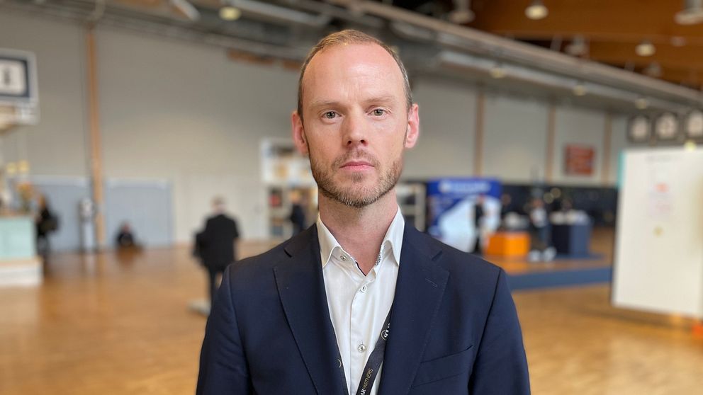 En person i kostym står i en konferenslokal med andra deltagare i bakgrunden.