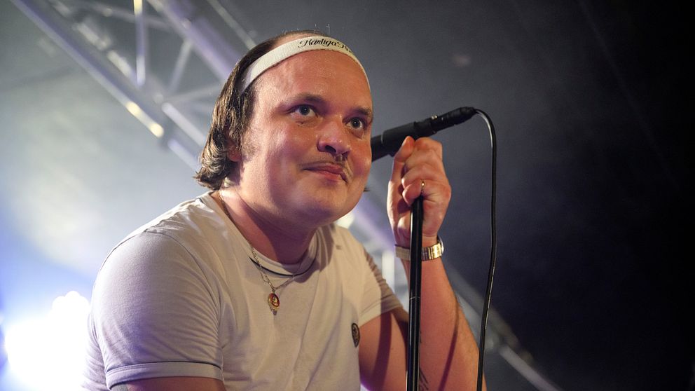 En manlig sångare i en vit t-shirt står vid en mikrofon med en scenbelysning som skapar en dramatisk atmosfär. Han har på sig en armbandsklocka och en halskedja, och hans arm visar tatueringar, vilket bidrar till den musikaliska stämningen.