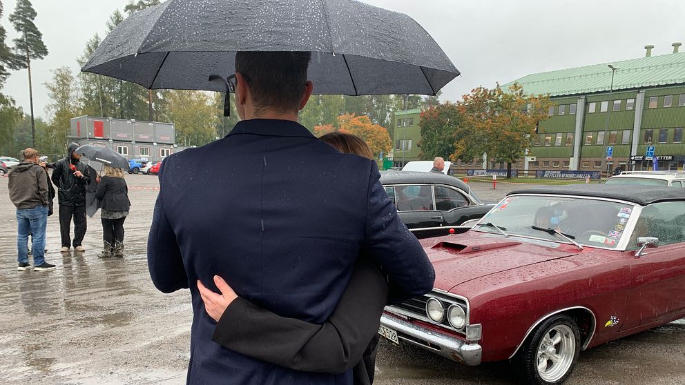 En man i en mörk kavaj håller om en kvinna under ett paraply, medan de står vid en röd veteranbil. Det regnar och flera personer syns i bakgrunden, som också är klädda för vädret. Området är en parkeringsplats omgiven av träd och byggnader.