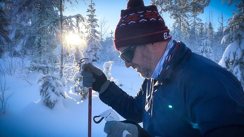 En man med mössa och svarta heltäckande glasögon åker skidor i en snötäckt skog