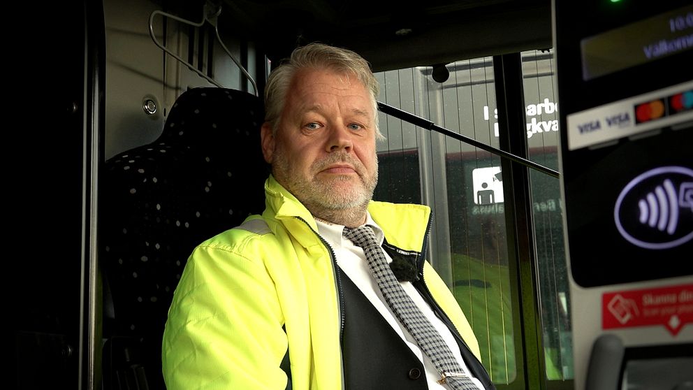 En person i en gula reflexjacka sitter vid ratten i en buss. Bakom syns bussens interiör och ett betalningssystem med kortläsare.
