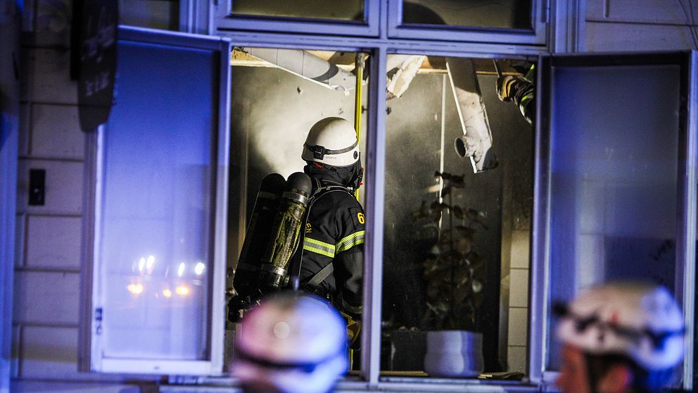 Brandman synlig genom fönster under släckningsarbete i byggnad. Två hjälmklädda personer i förgrunden.